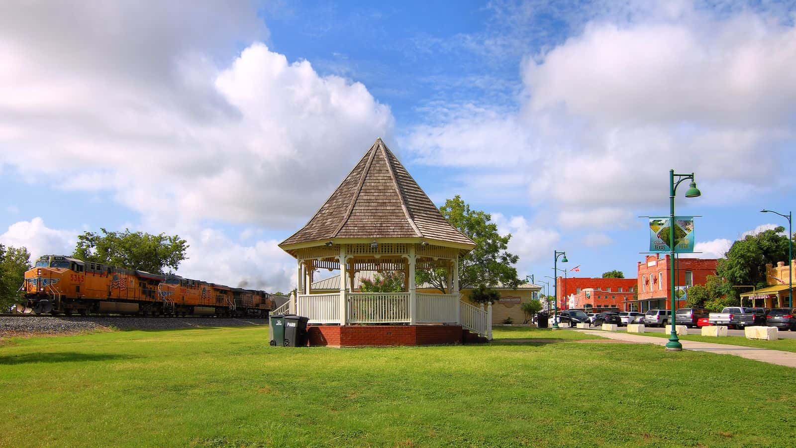Glorieta blanca sobre el césped en el centro de Buda, con un tren de carga detrás y edificios de ladrillo a la derecha