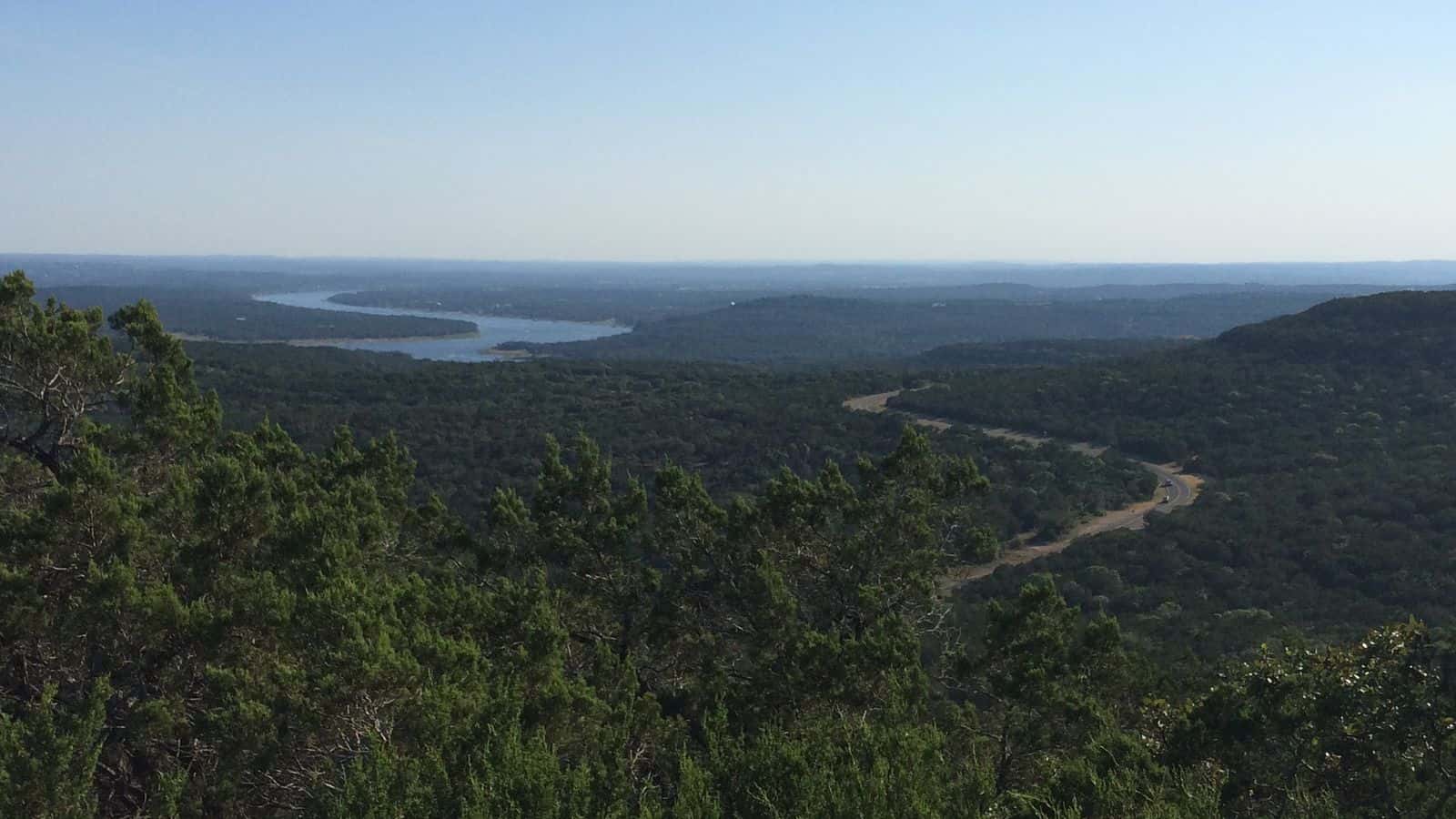 Lake Travis a lo lejos entre colinas cubiertas de árboles, con una carretera que serpentea por el valle, visto desde un mirador de Warbler Vista junto a Lago Vista
